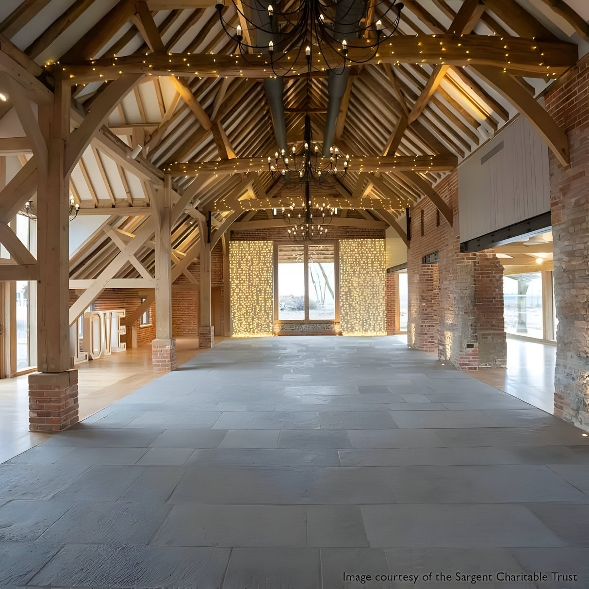 A spacious rustic barn interior features exposed wooden beams, brick walls, and large windows. The ceiling is adorned with the Carisbrooke 18 Light Chandelier - Black and string lights. A bright LOVE sign on the left enhances the romantic ambiance alongside candelabra-style arms.