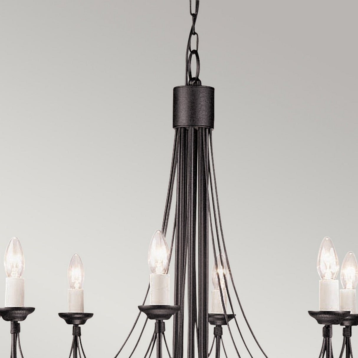 The Carisbrooke 8 Light Chandelier - Black showcases a vintage charm with its minimalist and sophisticated black metal design. With a Gothic inspiration, it includes multiple candle-shaped bulbs and elegantly curved arms, all suspended from a chain against a gray backdrop.