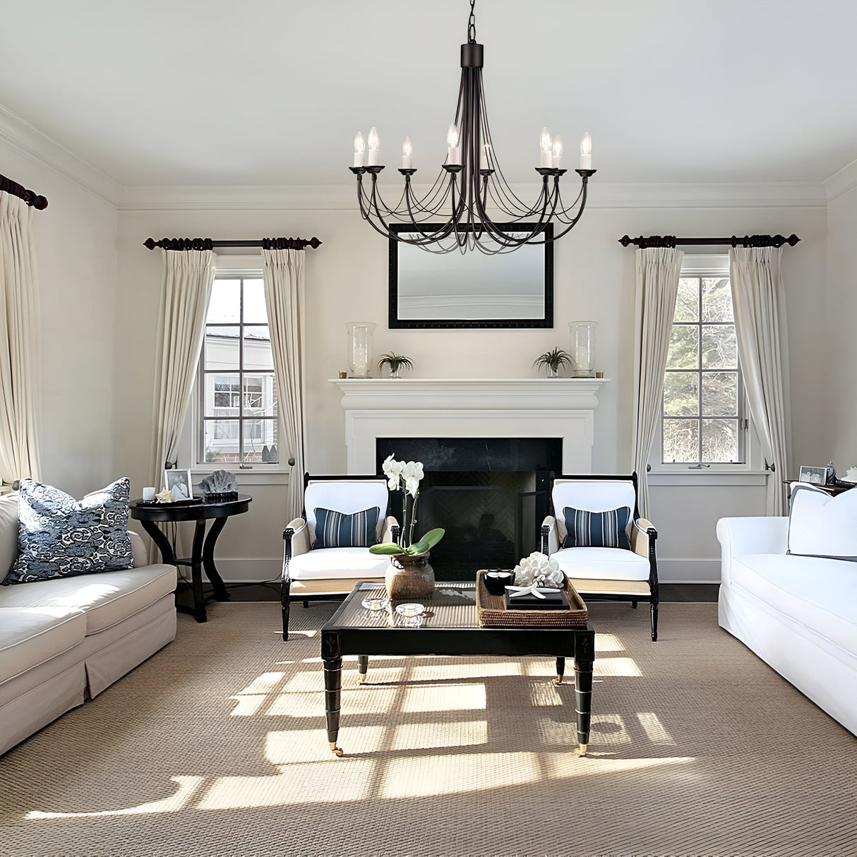 An elegant living room with a white color scheme features a black Carisbrooke 8 Light Chandelier and white sofas. A sleek black coffee table sits on a beige carpet, while the fireplace is adorned with a mirror and plants. Tall windows with white curtains let in natural light, adding vintage appeal.