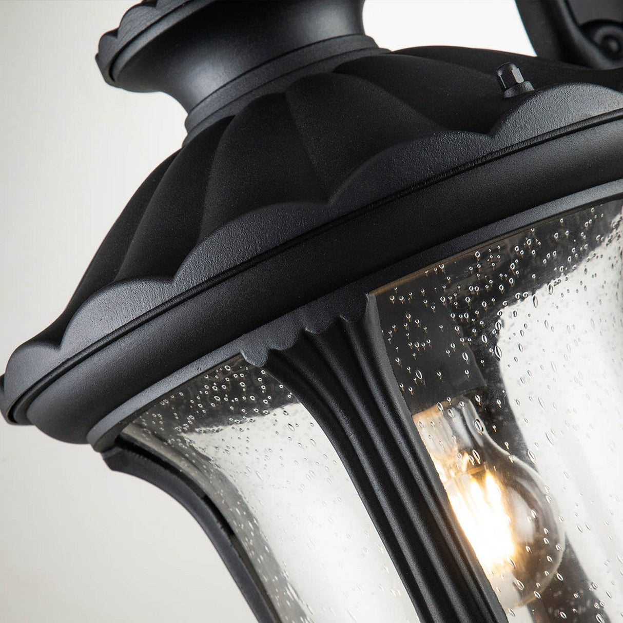 The Chicago Outdoor Medium Wall Lantern - Black, featuring a scalloped edge and bubbled glass panels, exudes vintage charm. Inside, a glowing bulb casts a warm light, making it an ideal outdoor lighting solution.