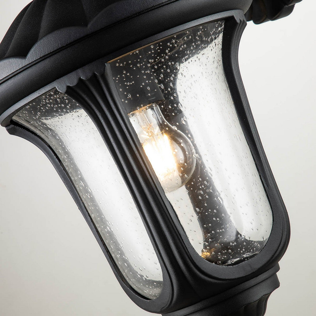 Close-up of the Chicago Outdoor Medium Wall Lantern - Black highlighting its vintage charm with a textured clear glass shade. The visible bulb inside emits a soft glow, accentuated by the water droplet design on the glass, making it an elegant outdoor lighting solution.