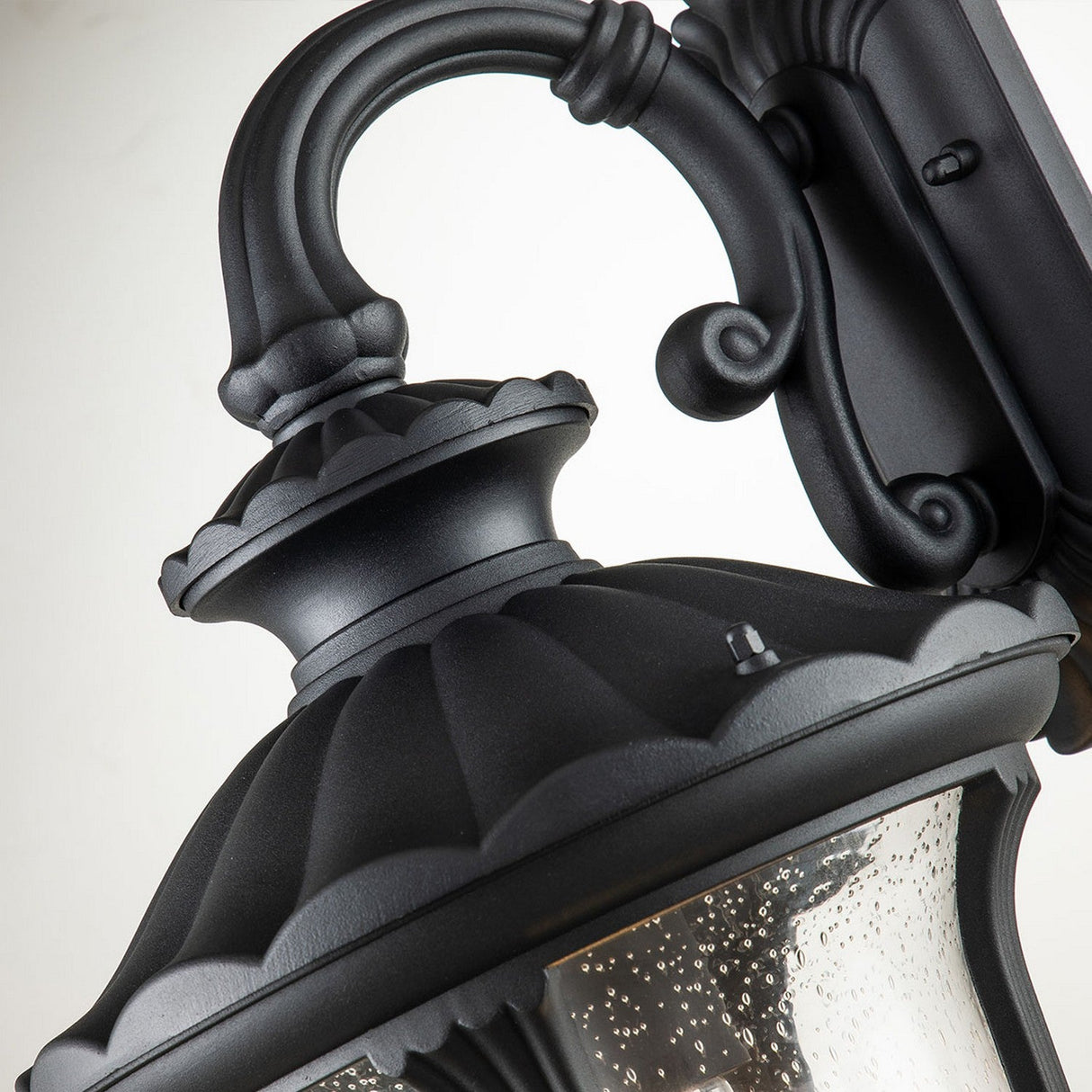 Close-up of the Chicago Outdoor Medium Wall Lantern - Black, a vintage-style outdoor lighting solution. The black metal fixture features ornate detailing with a curved arm and textured glass shade, seamlessly combining vintage charm with classic and elegant elements.