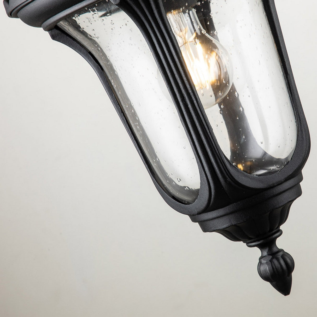 A close-up of the Chicago Outdoor Small Wall Lantern - Black exudes vintage elegance with its clear glass casing, adorned with raindrops. Illuminated by a glowing bulb inside, the durable aluminum frame stands out against a light gray background.