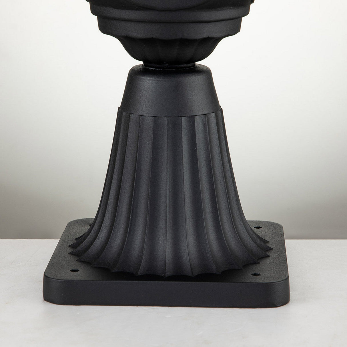 The Chicago Small Pedestal Lantern - Black features a vintage design with its black, fluted pedestal base and square bottom platform. The vertical grooves and slightly rounded middle section provide a smooth finish, making it ideal for supporting an outdoor lantern against a neutral backdrop.