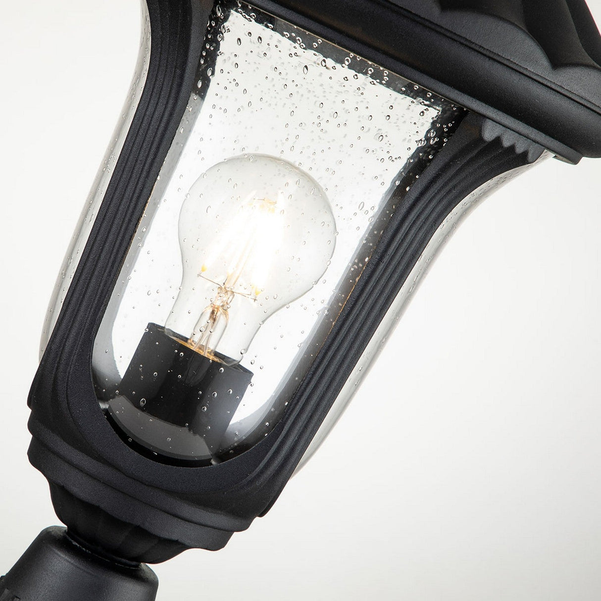 Close-up of the Chicago Small Pedestal Lantern in black, adorned with raindrops on its glass and featuring a vintage design. The illuminated bulb within casts a soft glow, highlighted against the plain white background to accentuate the elegance of this garden lighting piece.