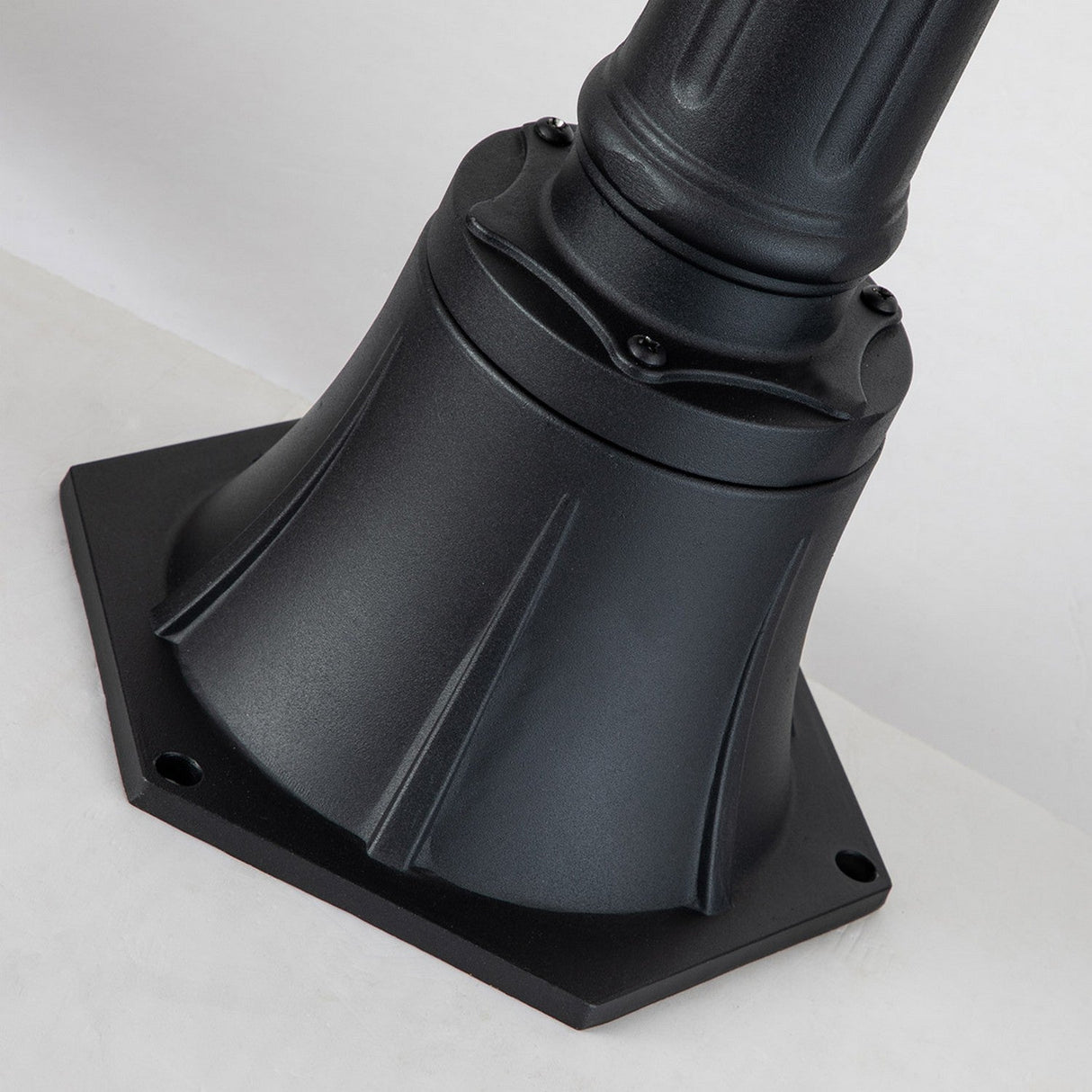 A detailed view of a stylish, black metal base with a hexagonal form and decorative ridges captures a vintage aesthetic. Firmly mounted on a flat white surface, it appears to support an outdoor lighting fixture, similar to the Chicago Small Pillar Lantern - Black.