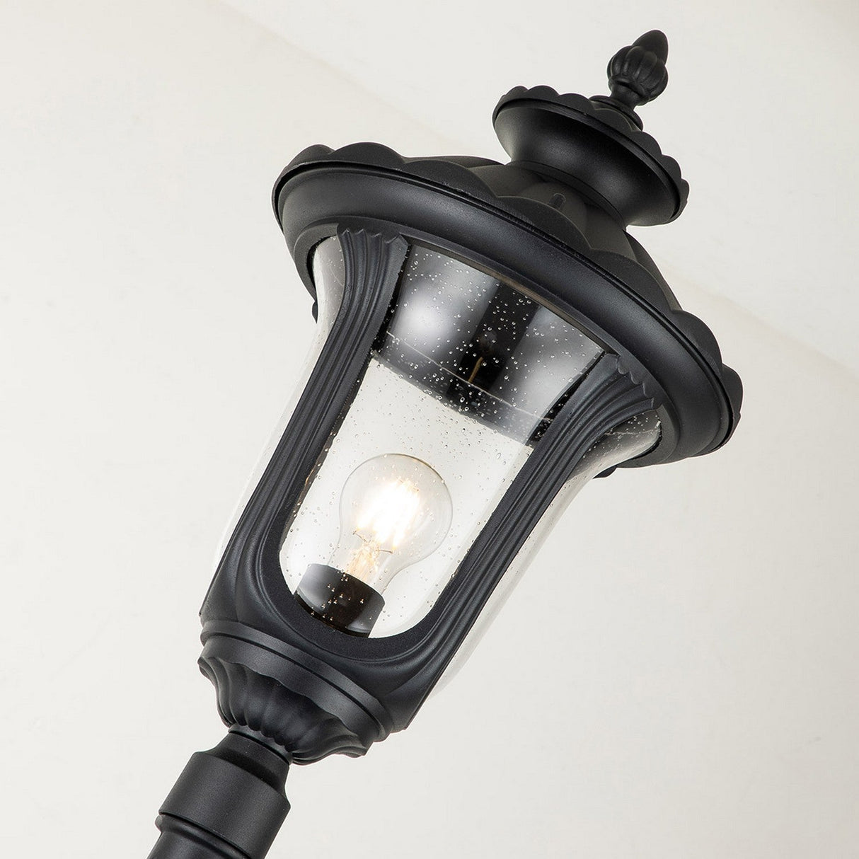 The Chicago Single Head Lamp Post in black showcases a glowing bulb encased within transparent, slightly speckled glass. This vintage outdoor fixture features decorative detailing and is captured from a low angle against a neutral background.