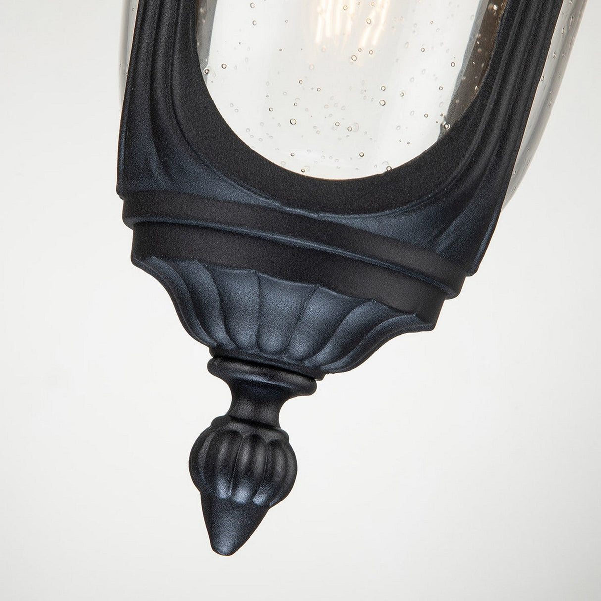 A close-up image features the Chicago Outdoor 1 Light Small Chain Pendant in black, showcasing its ornate and vintage charm. The fixture's textured design is highlighted in the clear seeded glass housing, with a curved metal base that enhances its classic appeal against a plain backdrop.