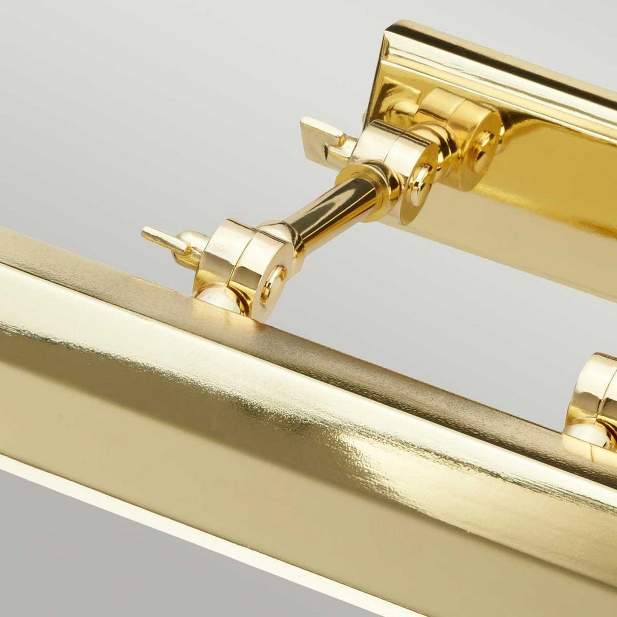 A close-up of a shiny hinge system in polished brass shows cylindrical pivot points and smooth, reflective surfaces. This hinge, featuring adjustable knuckle joints, is attached to a similar golden structure, offering a sleek and modern aesthetic reminiscent of the Chawton Large Picture Light in Polished Brass.
