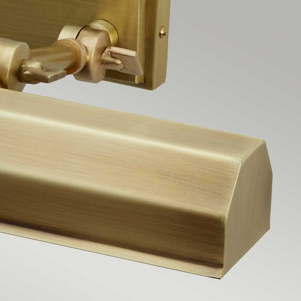 The Chawton Medium Picture Light in aged brass is showcased mounted on a wall. Its angular design features a brushed finish and adjustable knuckle joints for directing the light, set against a neutral gray background.