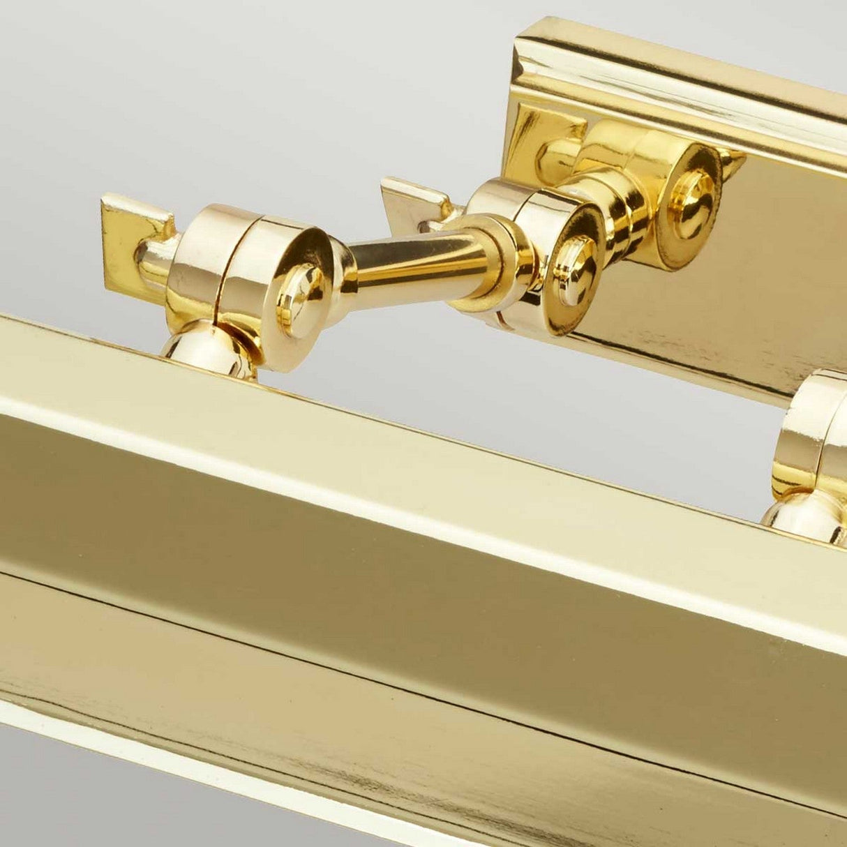 A close-up of the Chawton Medium Picture Light - Polished Brass highlights its two adjustable arms with knuckle joints and round hinges, set against a light-colored, neutral background that accentuates the shiny, reflective metal finish.
