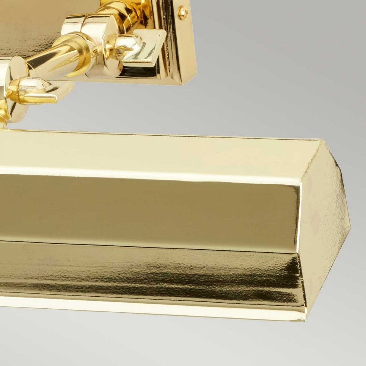 A detailed view of the Chawton Medium Picture Light - Polished Brass, showcasing its lustrous finish, rectangular shade, decorative features, and adjustable knuckle joints against a neutral gray backdrop.