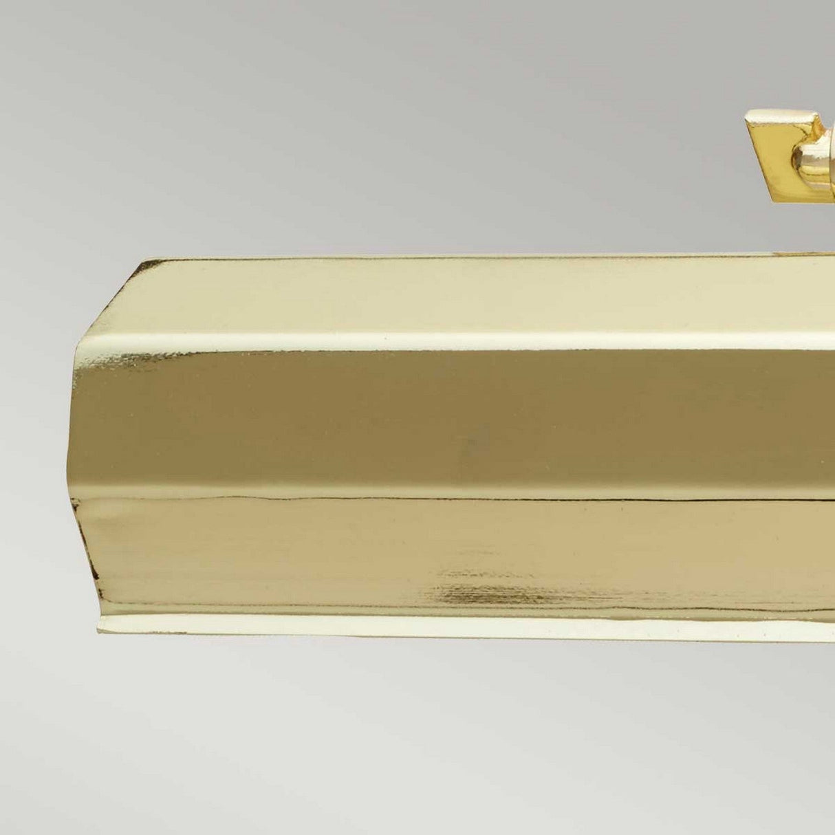 A close-up of the Chawton Medium Picture Light in polished brass showcases its smooth, reflective surface against a gray background. An adjustable knuckle joint is elegantly attached to the top right corner, reminiscent of classic designs.