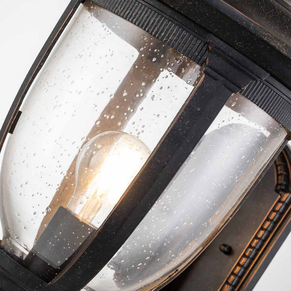 Close-up of the Cleveland-1 Outdoor Medium Wall Lantern, featuring clear seeded glass elegantly encasing a glowing bulb. The weathered bronze finish adds rustic charm to its design.