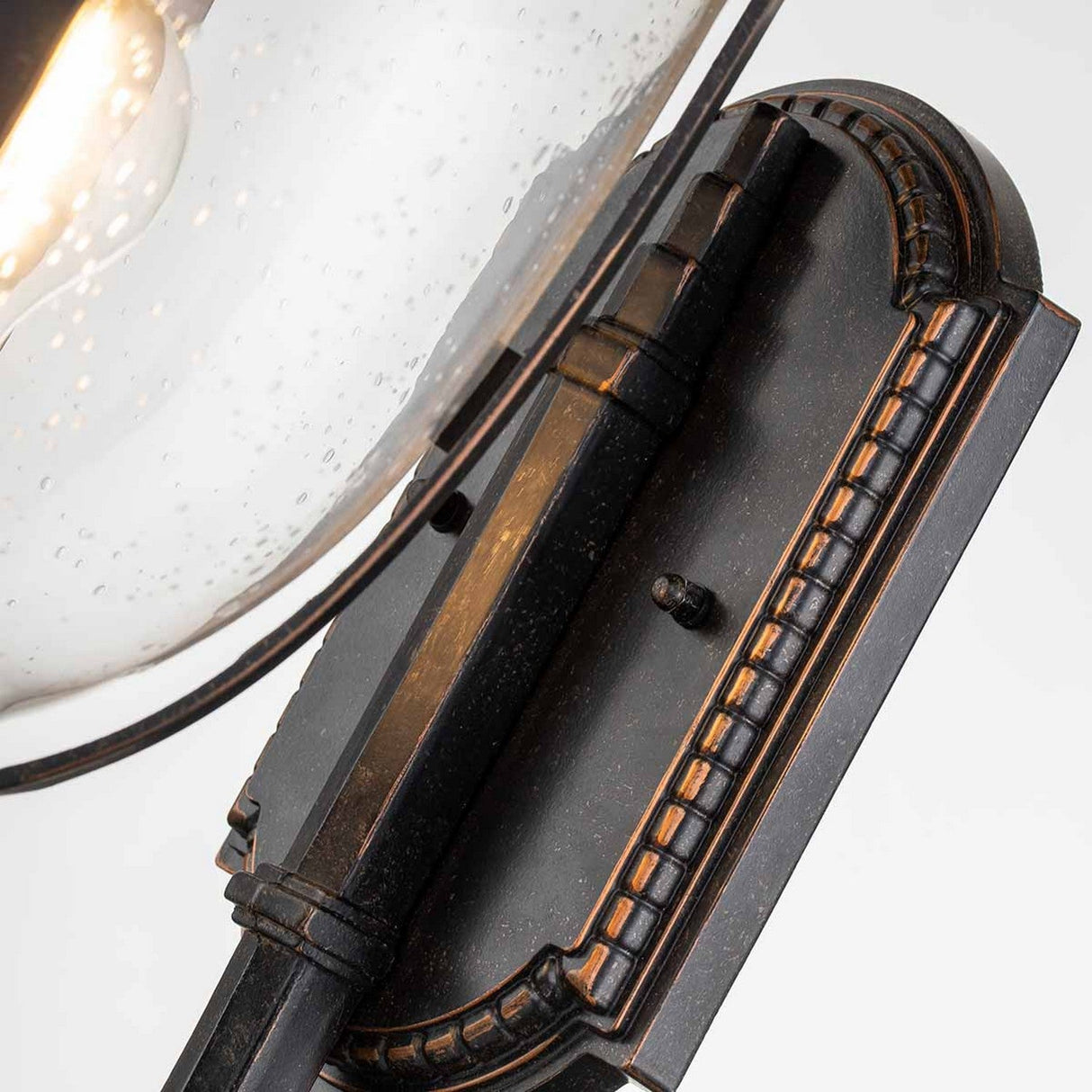 A close-up of the Cleveland-1 Outdoor Medium Wall Lantern in weathered bronze showcases its ornate detailing. The fixture includes a clear seeded glass shade that covers the light bulb, with delicate specks that enhance its elegant, vintage charm, making it ideal for an outdoor setting.