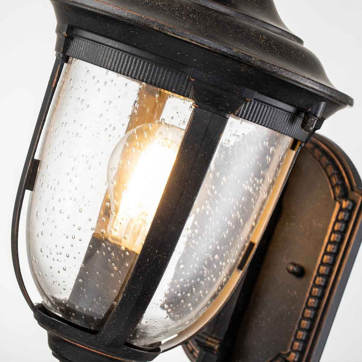 Close-up of the Cleveland-1 Outdoor Small Wall Lantern in weathered bronze, featuring clear seeded glass. A glowing bulb inside offers warm illumination, while the weathered bronze finish lends a rustic, vintage touch to its design.