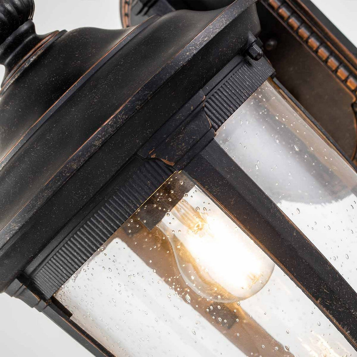 The Cleveland-2 Outdoor Medium Wall Lantern in a weathered bronze finish features a vintage-style design. Its glass is textured with small bubbles, encasing a lit bulb that emits a warm glow into outdoor areas.