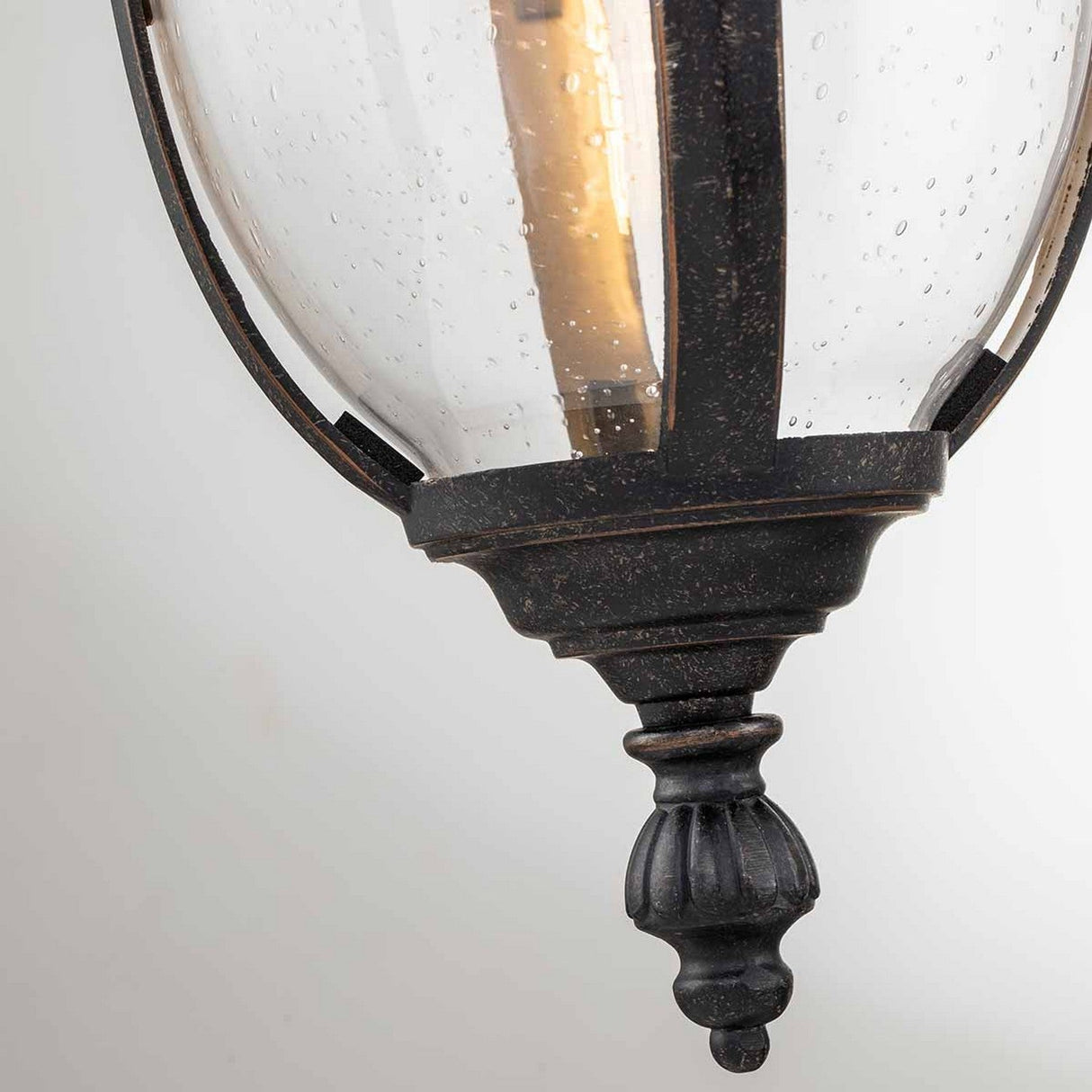 Close-up of the Cleveland-2 Outdoor Medium Wall Lantern in a vintage-style design, ideal for outdoor settings. The dark metal frame is finished in weathered bronze, complemented by clear glass panels. The bulb inside casts a warm glow, with small water droplets delicately adorning its surface.