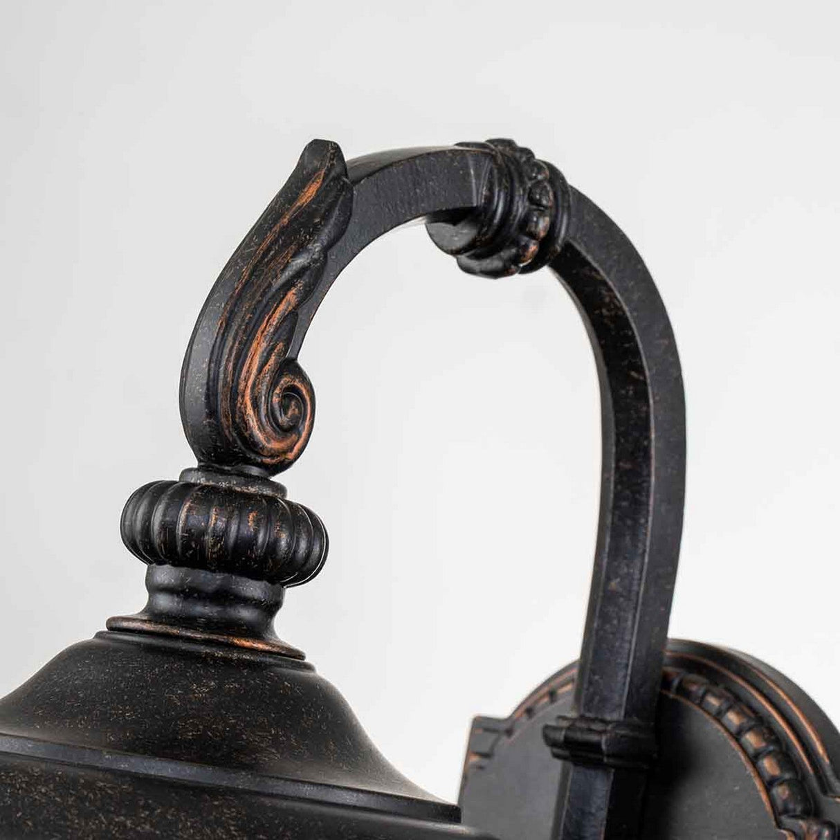 Close-up of the Cleveland-2 Outdoor Small Wall Lantern in weathered bronze, showcasing its vintage appeal with intricately detailed scrollwork on its curved arm, set against a plain white background.