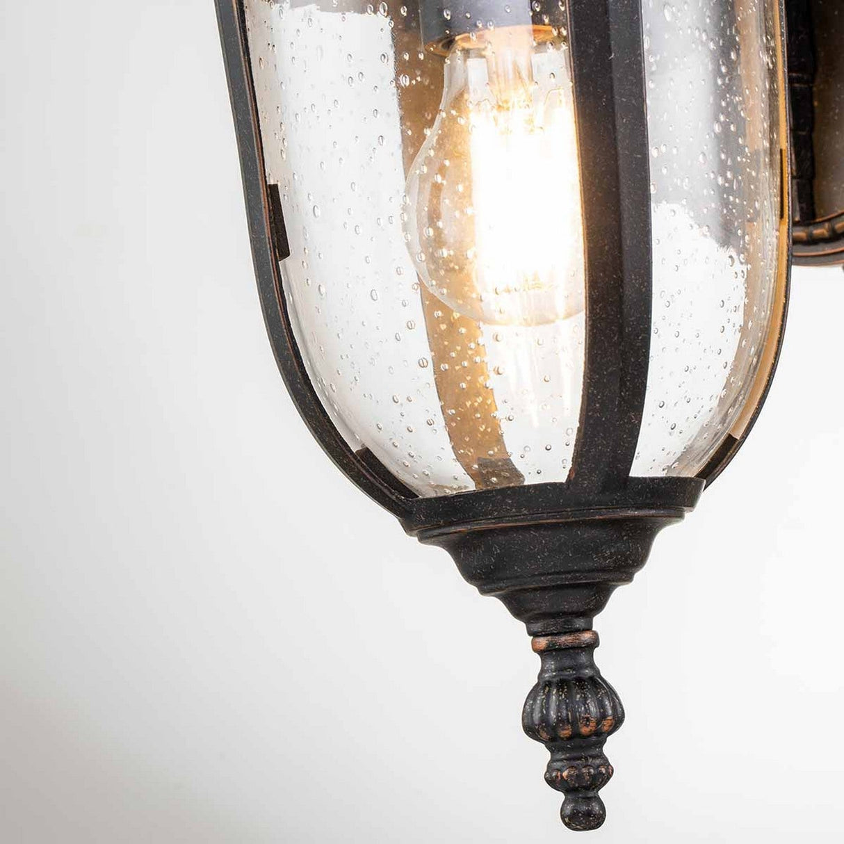 The Cleveland-2 Outdoor Small Wall Lantern showcases a close-up view featuring a clear, textured glass enclosure. Its illuminated bulb emits a warm glow, complemented by the rustic, weathered bronze finish of the metal frame. With an IP44 rating, it seamlessly blends style and functionality.