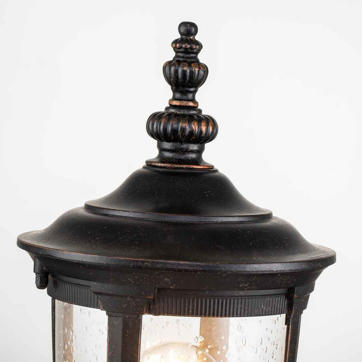 A detailed view of the Cleveland Small Pedestal Lantern in a weathered bronze finish, showcasing its vintage style with a black metal top adorned with decorative ridges. The slightly textured glass panels reveal a bulb inside, providing outdoor lighting with gentle illumination.