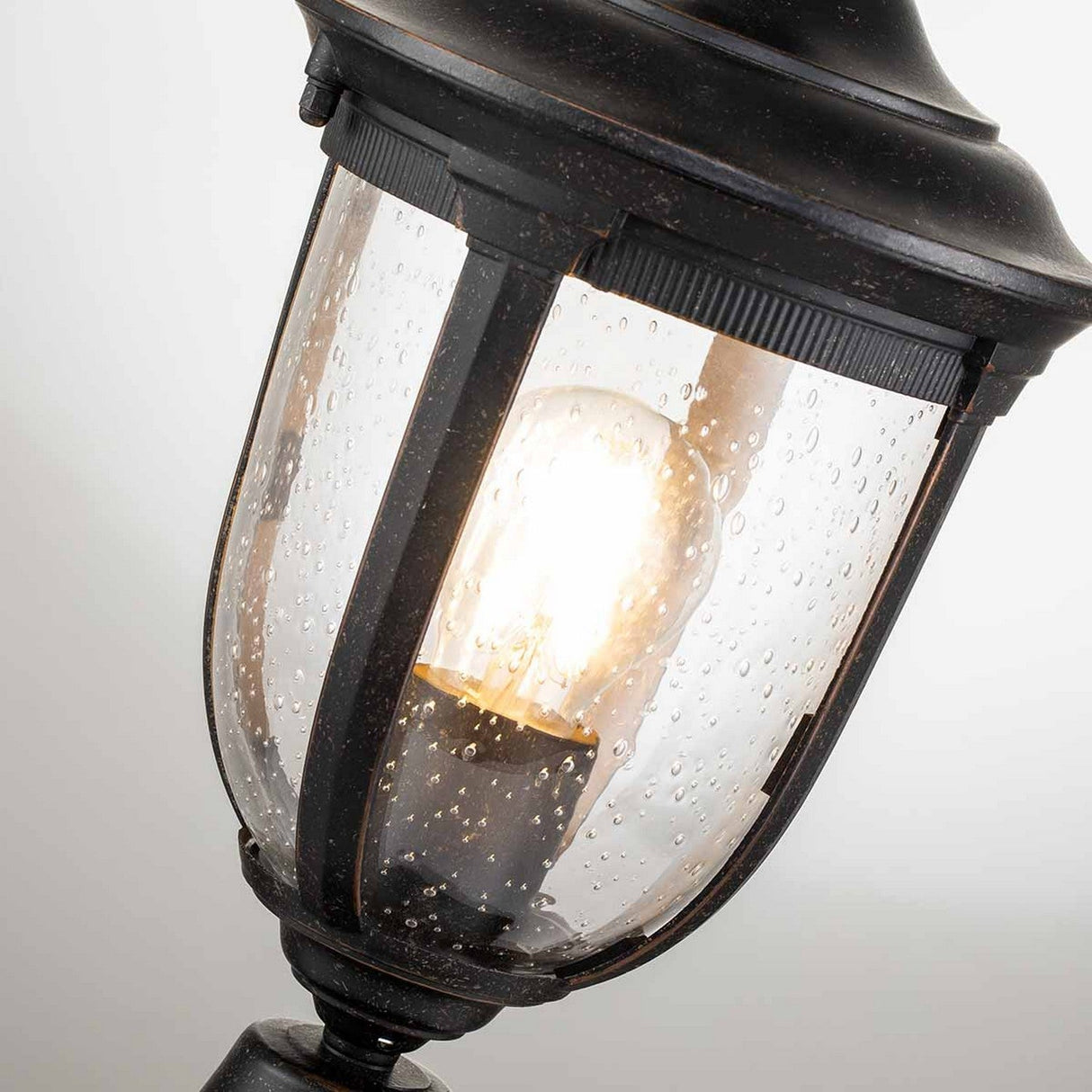 A close-up of the Cleveland Small Pedestal Lantern showcases its vintage design. The lantern's clear glass is adorned with water droplets, revealing a glowing filament bulb inside. The aluminum frame features a weathered bronze finish, providing a classic and rustic look.