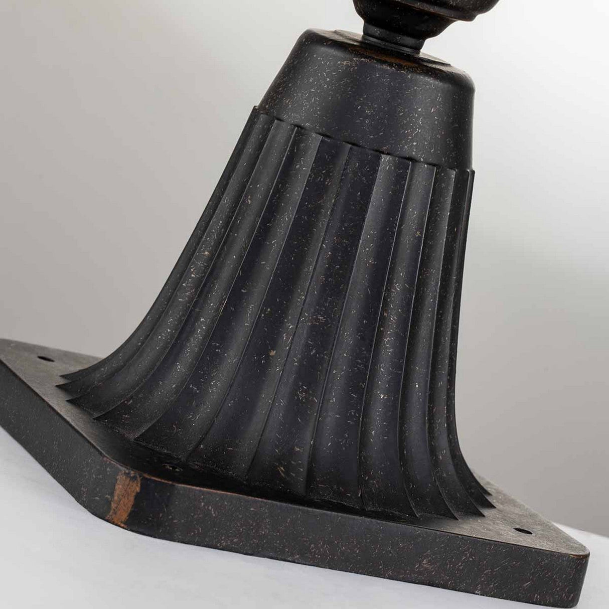 A close-up of the Cleveland Small Pedestal Lantern's fluted, dark metal base reveals a rectangular mounting plate. The weathered bronze finish displays a speckled texture with subtle hints of wear and age against a soft gray background.