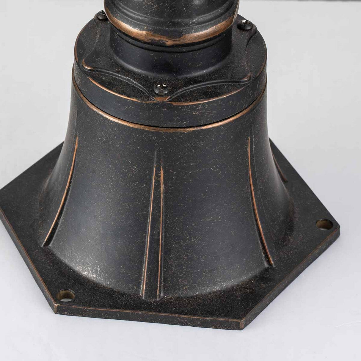 A close-up of the Cleveland Small Pillar Lantern reveals a dark metal pedestal base with a flared design and hexagonal bottom, finished in weathered bronze. The surface showcases visible screws and subtle distressed details, enhancing its vintage industrial style, making it perfect for outdoor lighting displays.