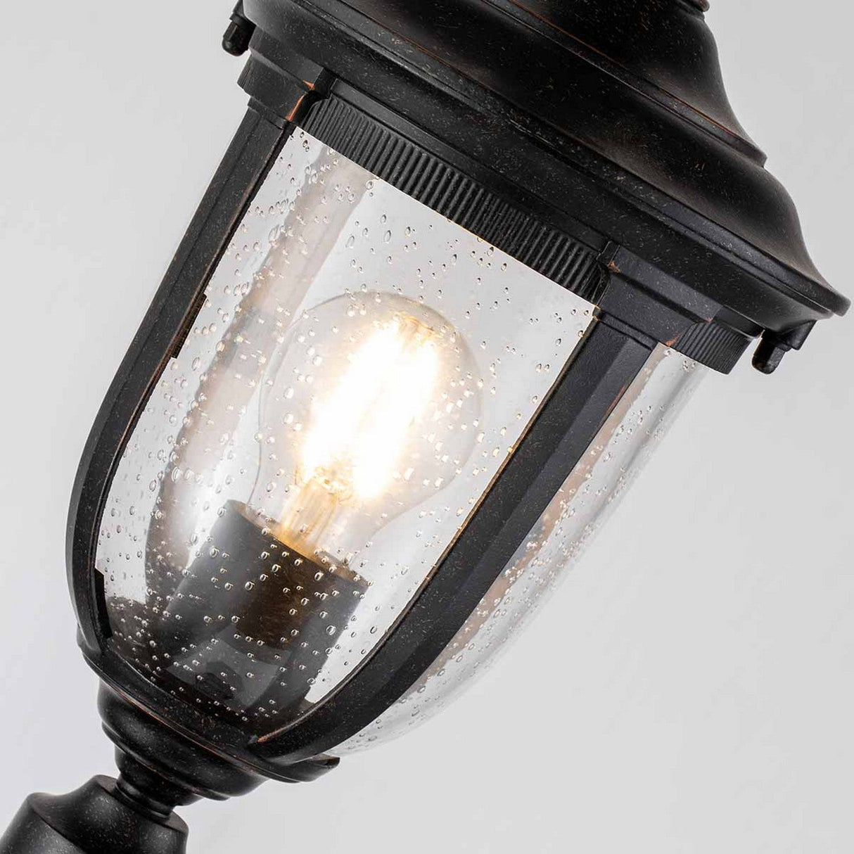 Close-up of the Cleveland Small Pillar Lantern in weathered bronze, showcasing its glowing bulb. The outdoor fixture is designed with a black metal frame and clear glass panels embellished with droplets for a textured appearance.