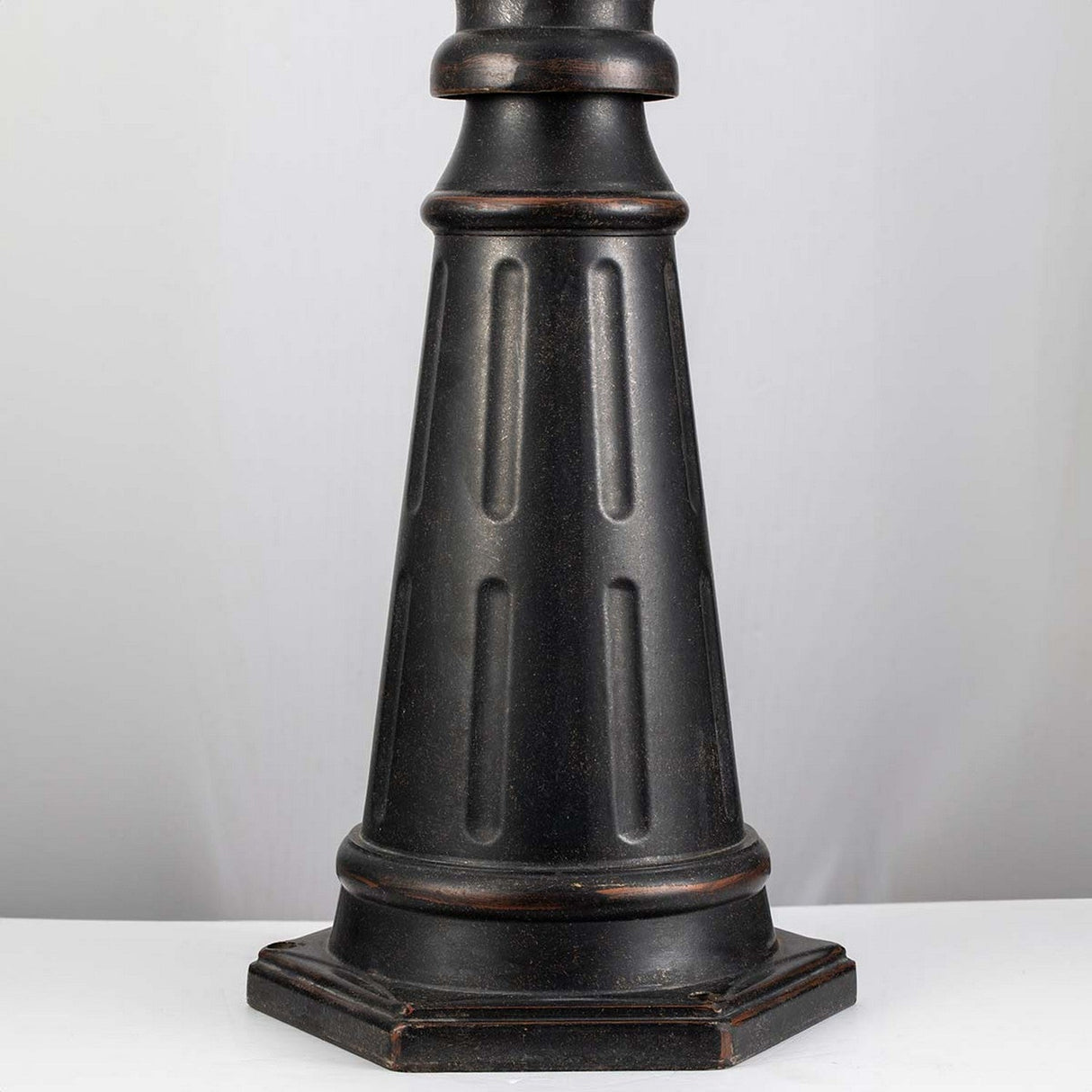 Standing against a plain white background, this tall, weathered bronze metal tripod leg features a tapered, cylindrical shape with vertical grooves and is set on a hexagonal base. It echoes the classic design of the Cleveland Single Head Lamp Post - Weathered Bronze created for outdoor space lighting.