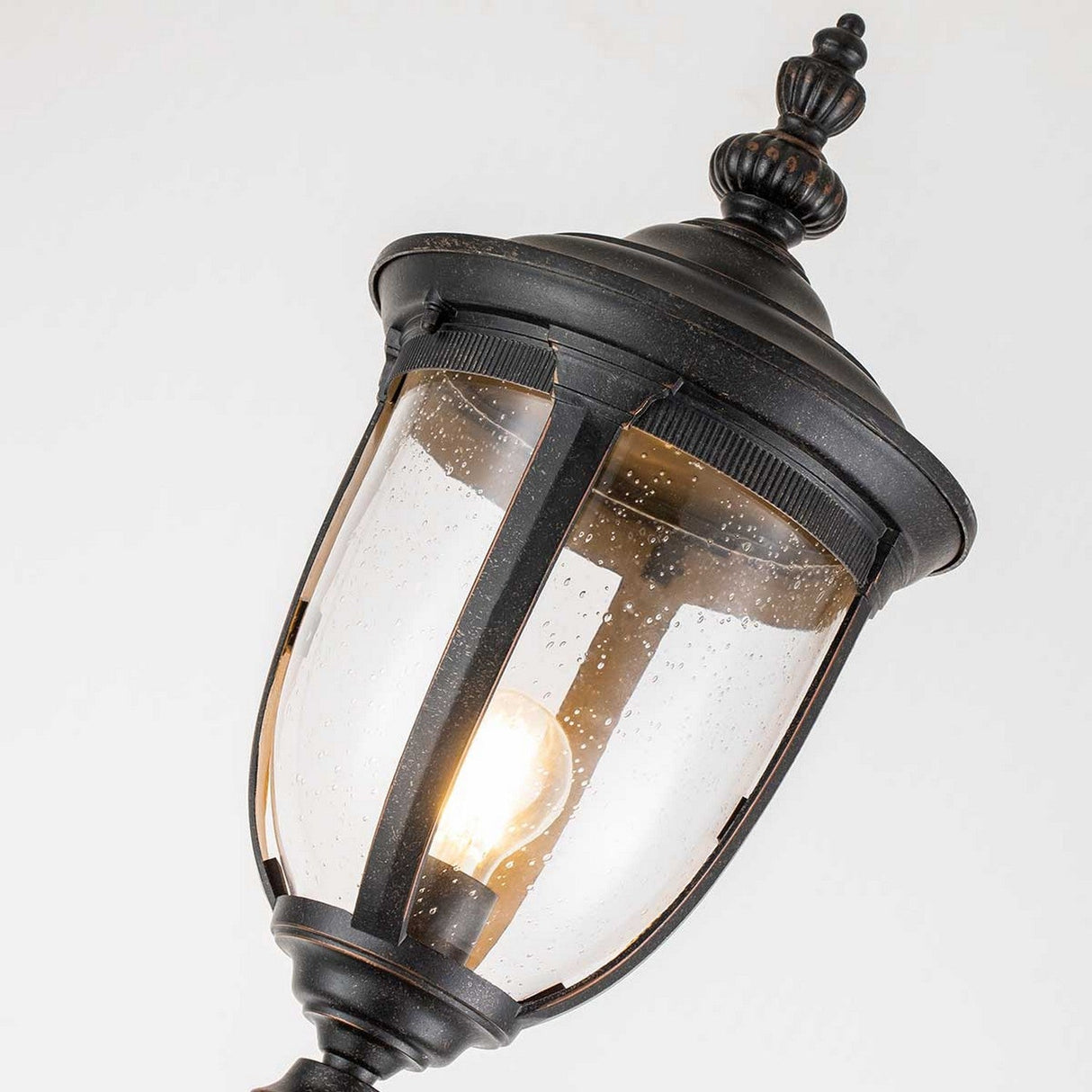 A close-up of the Cleveland Single Head Lamp Post - Weathered Bronze showcases its vintage-style black design with a decorative top and clear glass panels. Inside, an illuminated light bulb casts a warm glow, while the slightly speckled glass surface enhances its weathered bronze finish for an antique appearance.