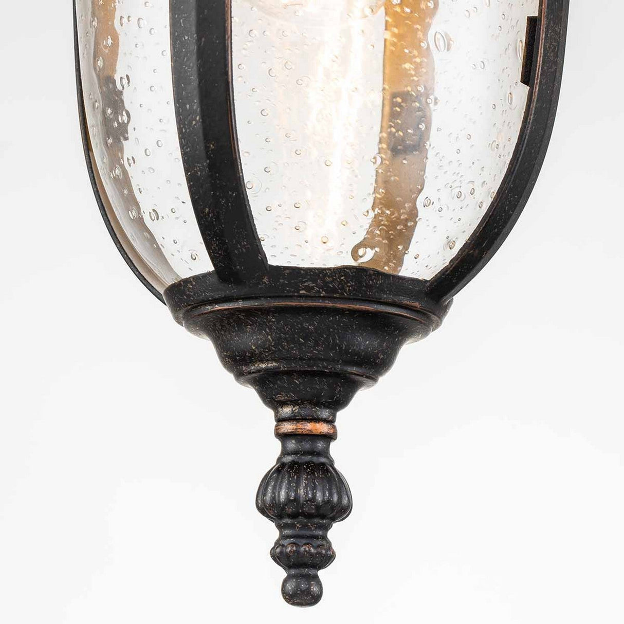A close-up of an outdoor lantern featuring a weathered bronze metal frame and clear, textured glass. The light inside emits a warm glow, embodying an ornate, vintage-style design reminiscent of the Cleveland Outdoor 1 Light Small Chain Pendant.