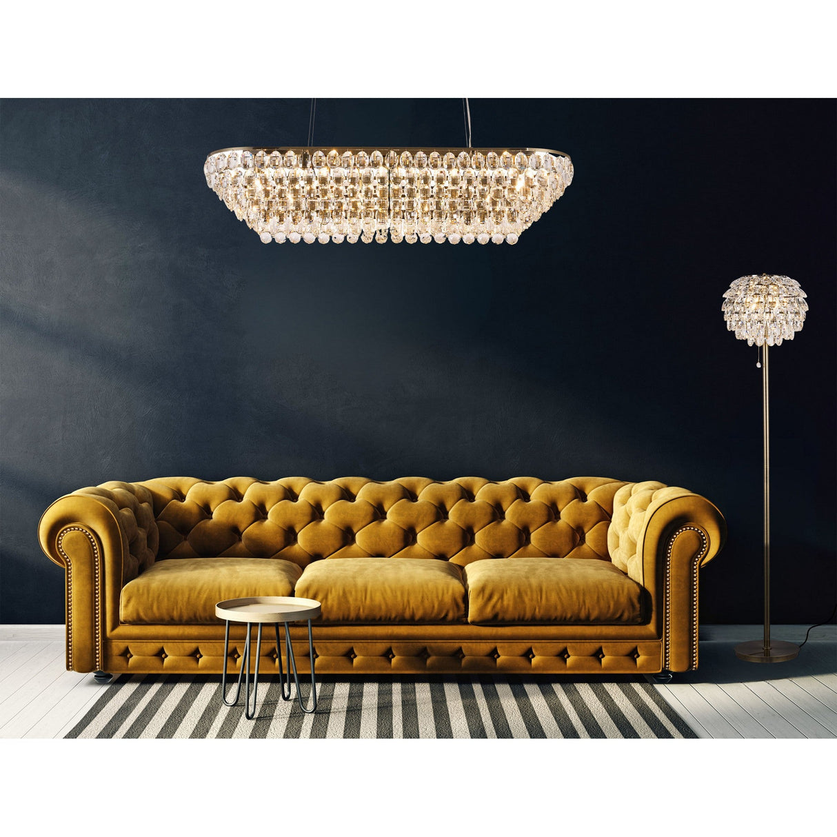 A stylish living room with a mustard-yellow velvet tufted sofa, striped rug, round coffee table, and crystal chandelier pairs beautifully with the Coniston 3 Light Crystal Floor Lamp in Antique Brass against a dark navy wall.