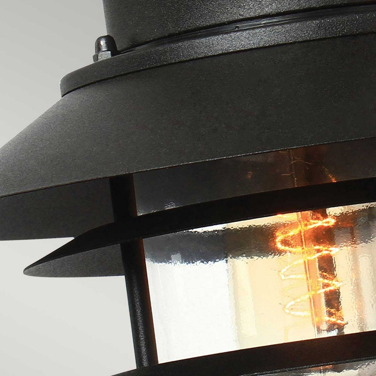 Close-up of the Copenhagen Outdoor Wall Lantern - Black, expertly constructed from black metal with a clear cylindrical glass housing that highlights a glowing filament bulb. This modern industrial design includes visible screws and a tiered shade, enhanced by stainless steel accents and an IP44 rating for enhanced durability.
