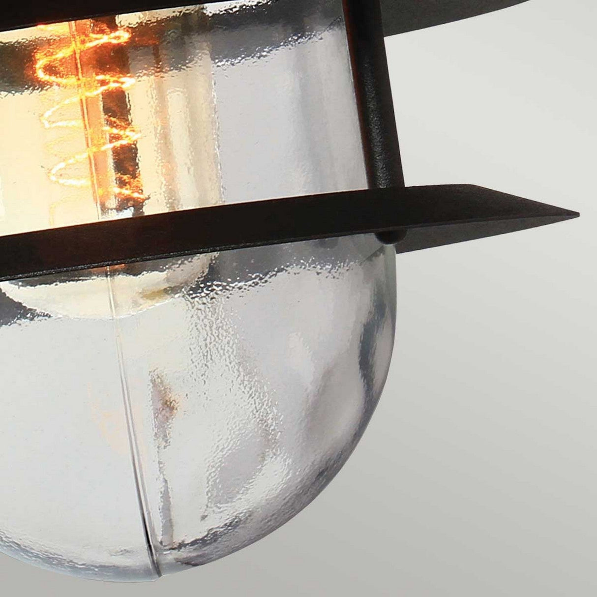 Close-up of a ceiling light fixture with a stainless steel frame and clear glass shade, showcasing a visible filament bulb that emits a warm glow. This modern piece exudes an industrial aesthetic, reminiscent of the sleek design found in the Copenhagen Outdoor Wall Lantern - Black, set against a neutral background.
