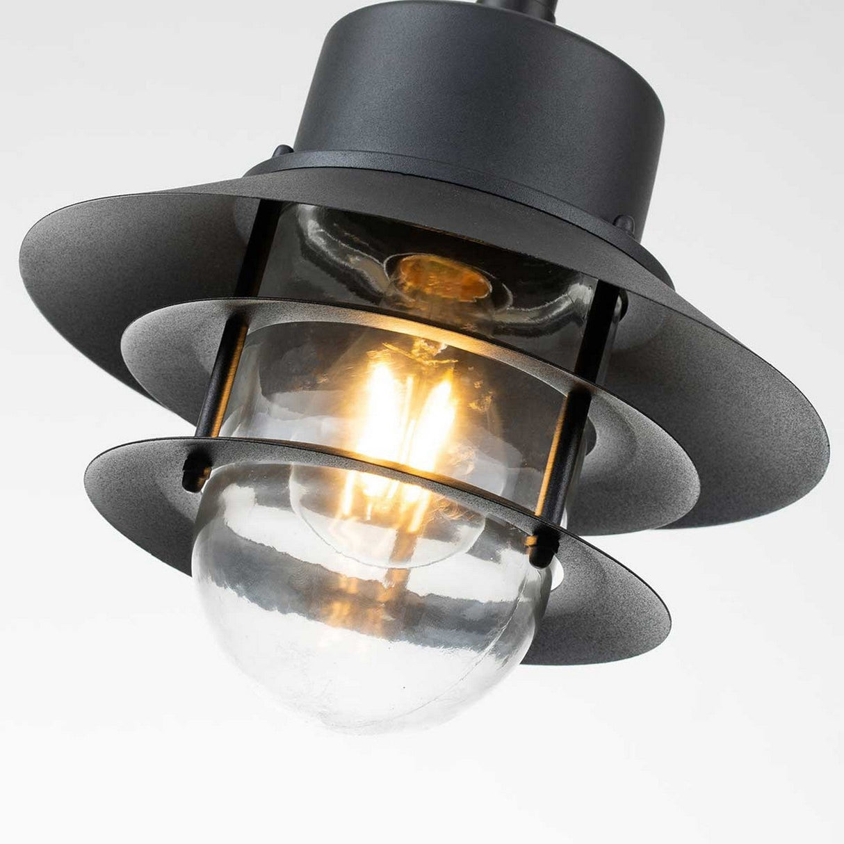 The Copenhagen Outdoor 1 Light Chain Pendant in black features a layered metal design and a clear glass cover that reveals the illuminated bulb inside, all set against a plain white backdrop, perfectly merging modern aesthetics with the simplicity of Danish design.