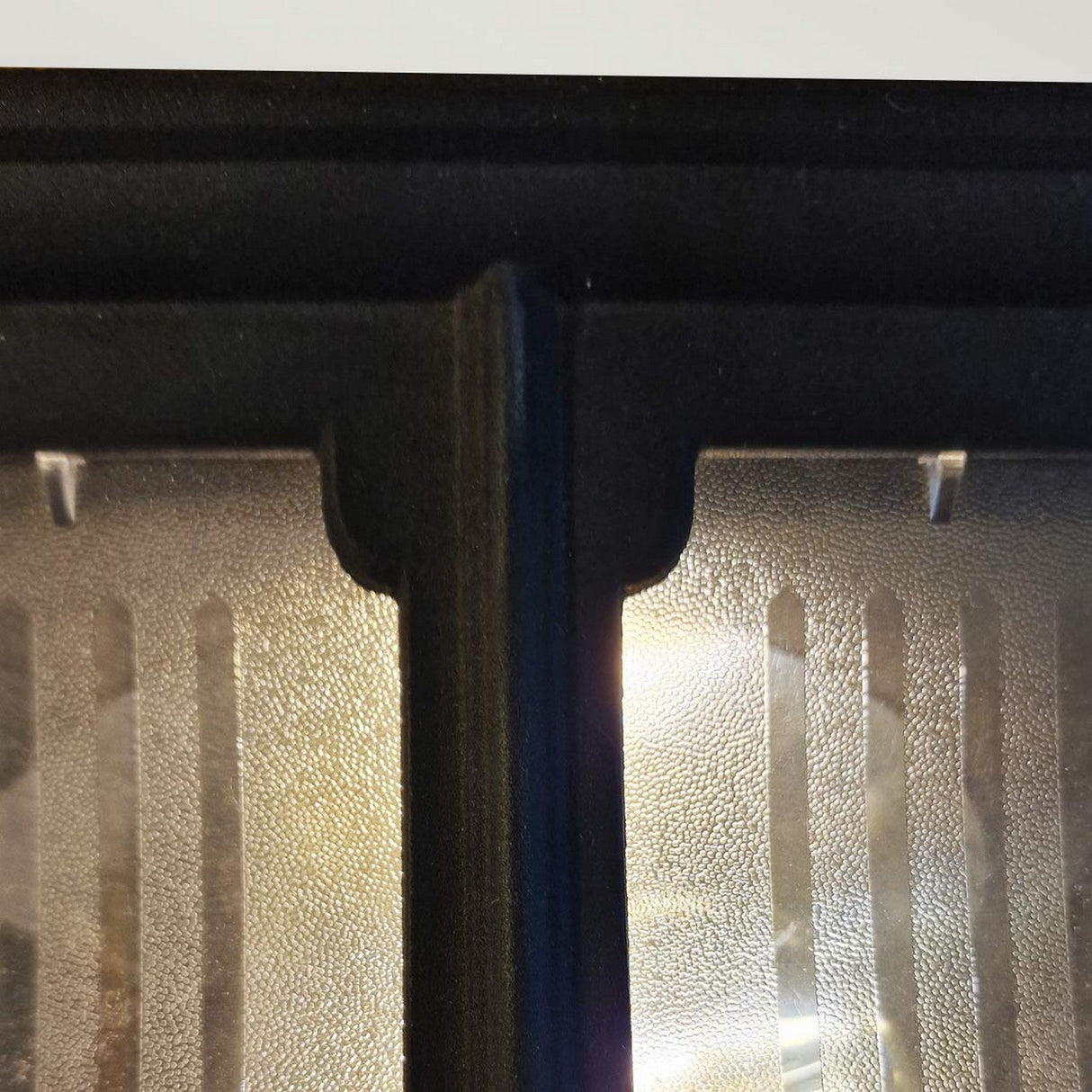 A close-up view reveals the textured glass surface of the Chapel 1 Light Outdoor Flush Light - Black, segmented by a black metal frame. Illuminated from behind, it casts captivating patterns on the glass, while its die-cast aluminum construction offers durability and an IP44 rating for outdoor use.