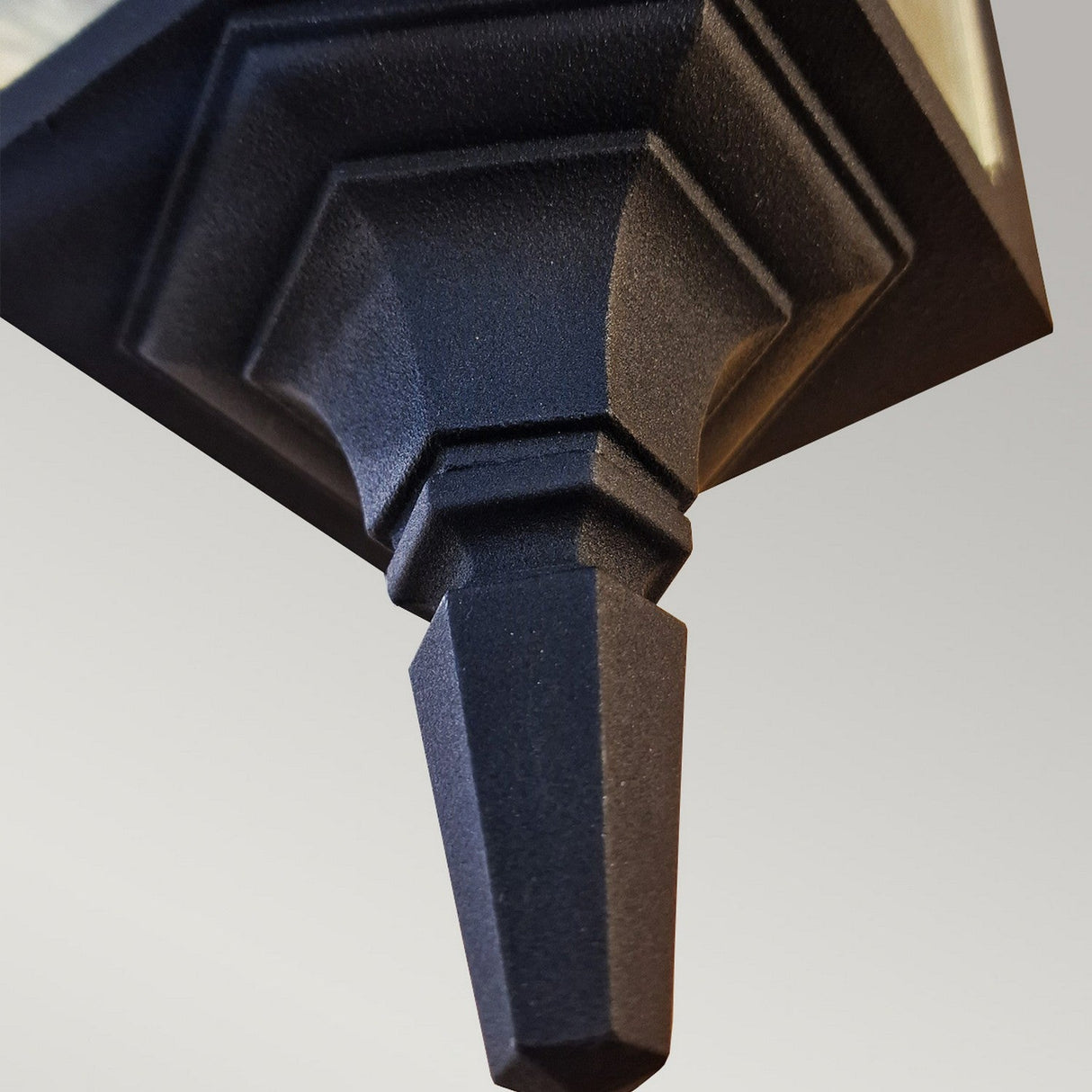 Close-up of the Chapel 1 Light Outdoor Down Wall Lantern in black, featuring a geometric, pointed structure extending from a larger form that resembles the tip of an outdoor wall light. The sleek black finish beautifully contrasts against the plain, light gray background, enhancing every detail of this striking piece with its IP44 rating assurance.
