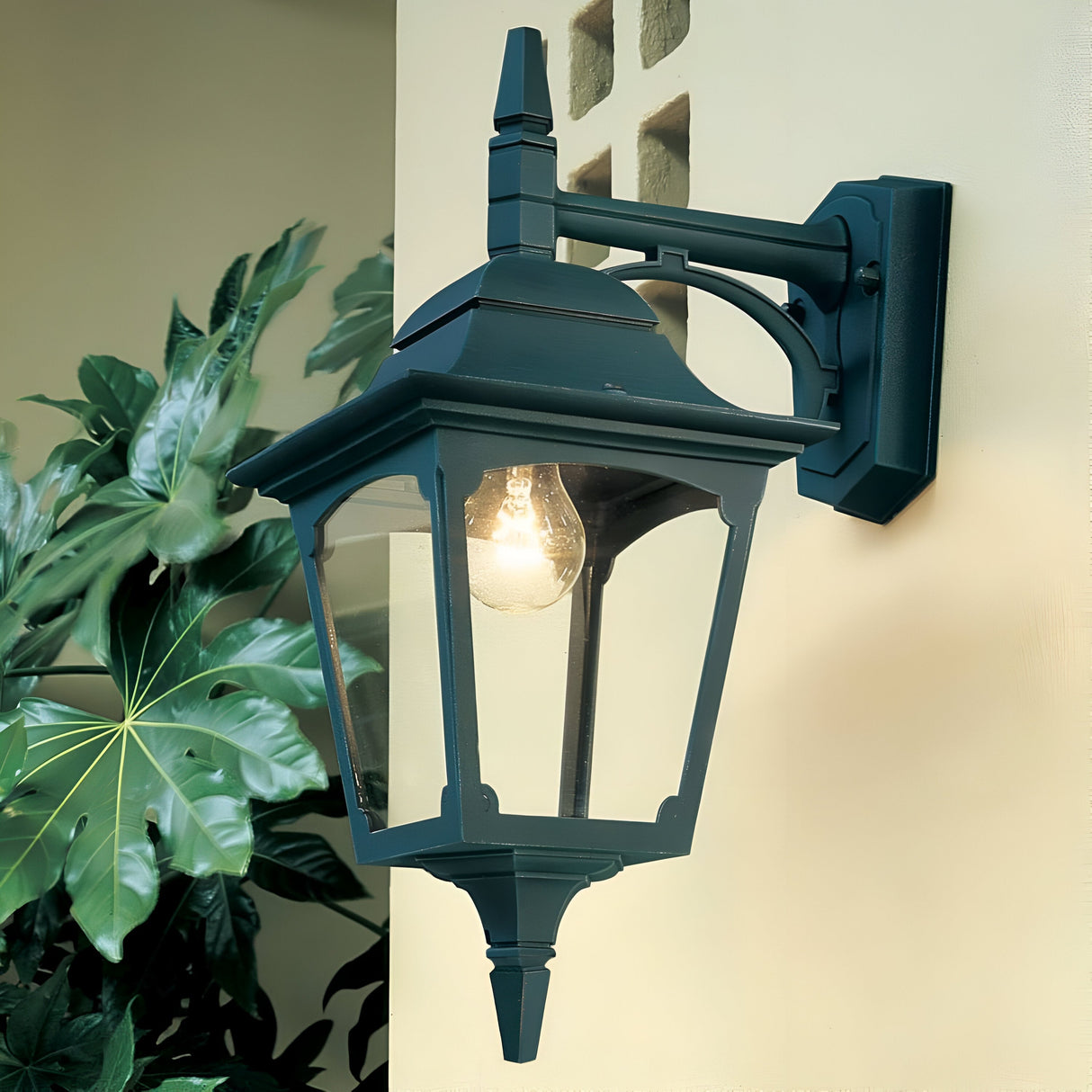 A Chapel 1 Light Outdoor Down Wall Lantern - Black, showcasing an IP44 rating and a clear glass enclosure that reveals the light bulb, is mounted on a beige wall. Large green leaves in the background enhance the natural elegance of this outdoor wall light setting.