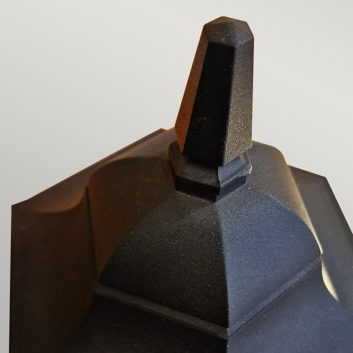 A close-up view of the Chapel 1 Light Outdoor Pedestal Lantern in black, showcasing its angular design with a pointed top, geometric shapes, and flat surfaces made from durable die-cast aluminum, set against a light background.