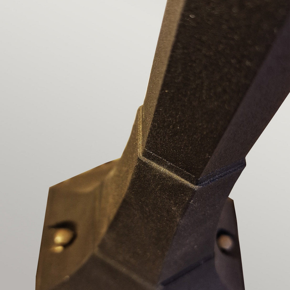 Close-up of the Chapel 1 Light Pillar Lantern - Black, crafted from die-cast aluminium featuring a textured surface and geometric design. It is securely attached to a base with visible screws. Ideal for outdoor lighting, its IP44 rating ensures it withstands the elements. The background is a neutral gray.