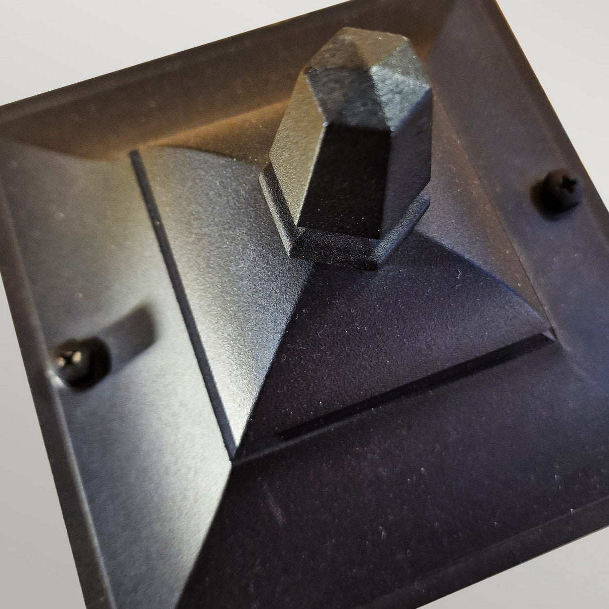 Close-up of the Chapel 1 Light Pillar Lantern - Black, showcasing its geometric design made from die-cast aluminum. The lantern features a pyramid-shaped top and two visible screw holes. Set against a plain background, it appears solid and angular in design, ready for seamless integration with outdoor lighting solutions.