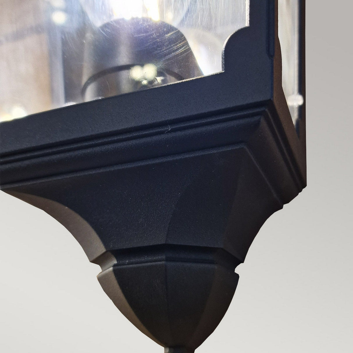 Close-up of the Chapel-1 1 Light Outdoor Wall Lantern in black, showcasing a die-cast aluminium lantern with a glass enclosure and a visible light bulb inside. The lantern, mounted against a light gray background, boasts a classic angular design and an IP44 weather resistance rating.