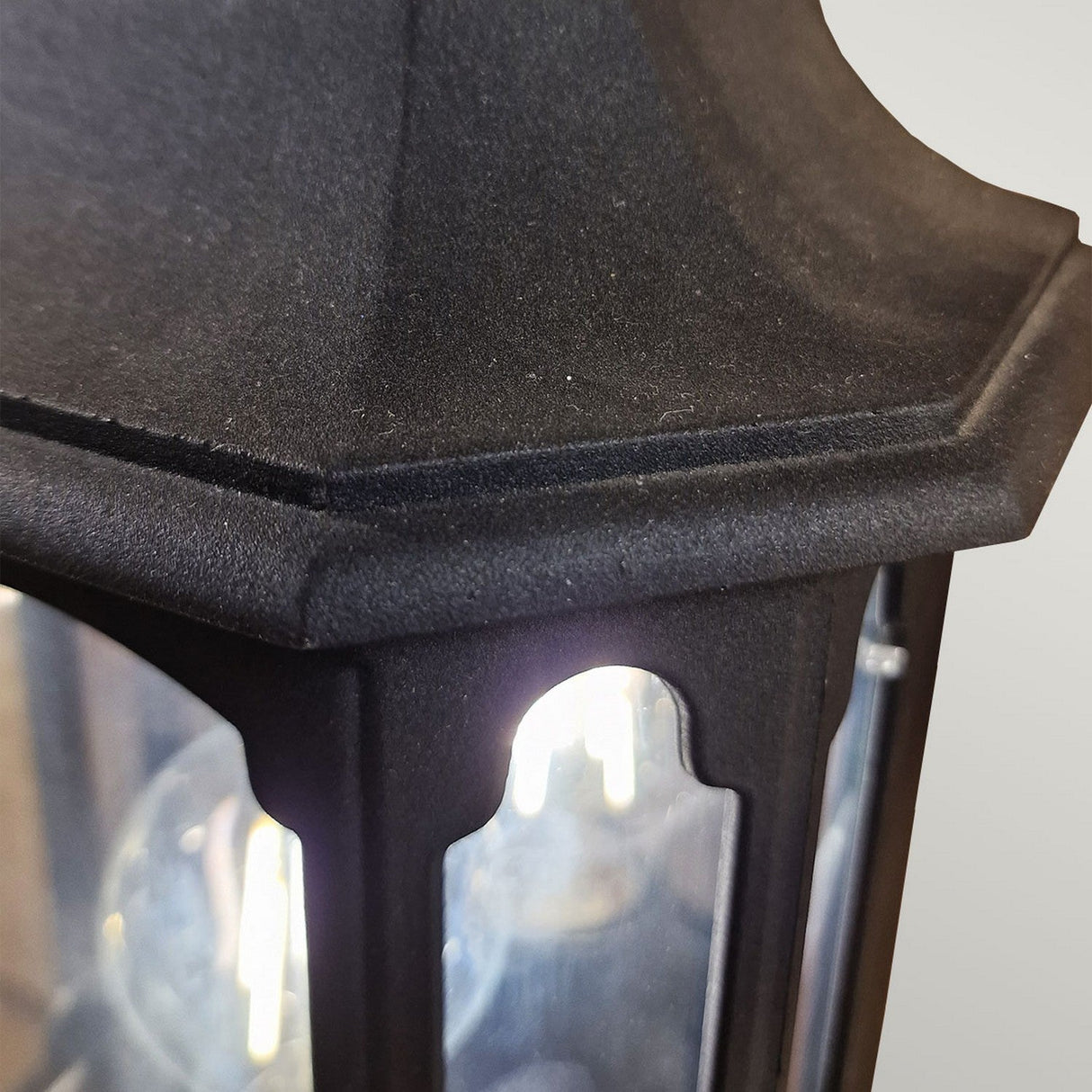 A close-up of the Chapel-2 1 Light Outdoor Wall Lantern in black, showcasing its glowing bulb within. This lantern is crafted from die-cast aluminum and features a textured surface with an angular design. The light emits a soft, warm glow that is partially visible through its glass panels.