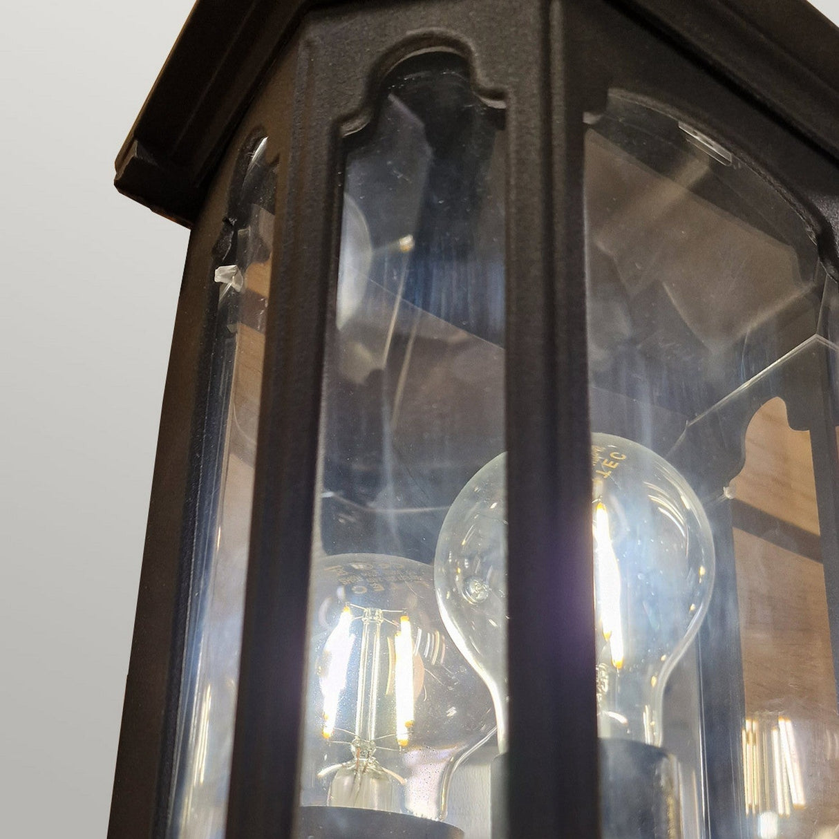A close-up view of the Chapel-2 1 Light Outdoor Wall Lantern in black showcases its lantern style and clear glass panels, crafted from sturdy die-cast aluminum. Inside, a glowing light bulb illuminates against a soft gray background.