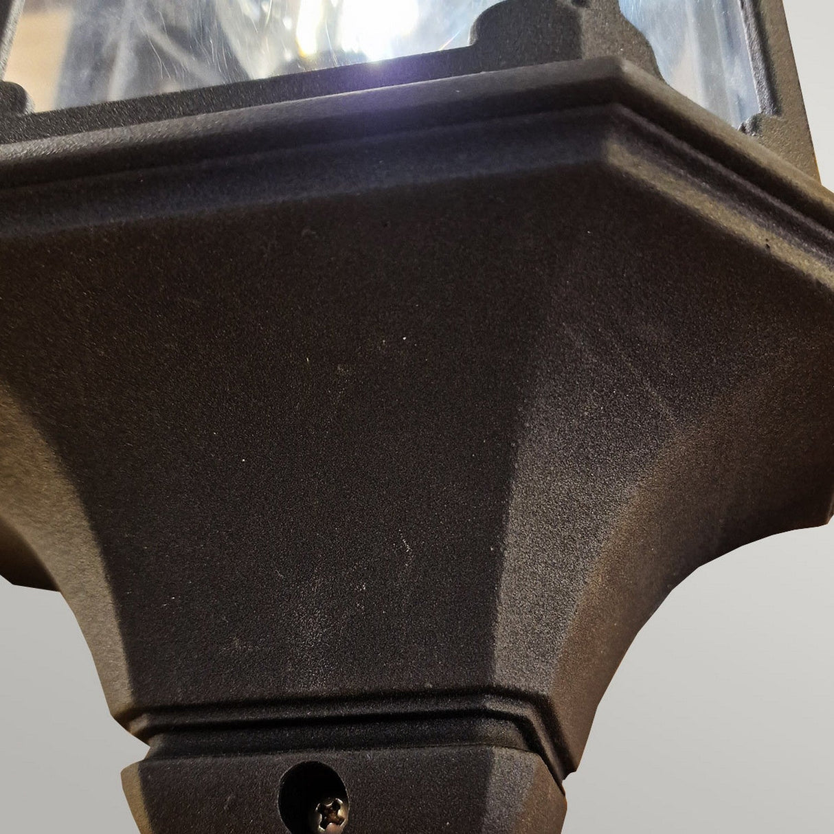 The Chapel-2 1 Light Outdoor Wall Lantern in black features a close-up of its die-cast aluminum base set against a light gray background. The top edge showcases a hint of reflective glass, making it ideal for outdoor wall lighting.