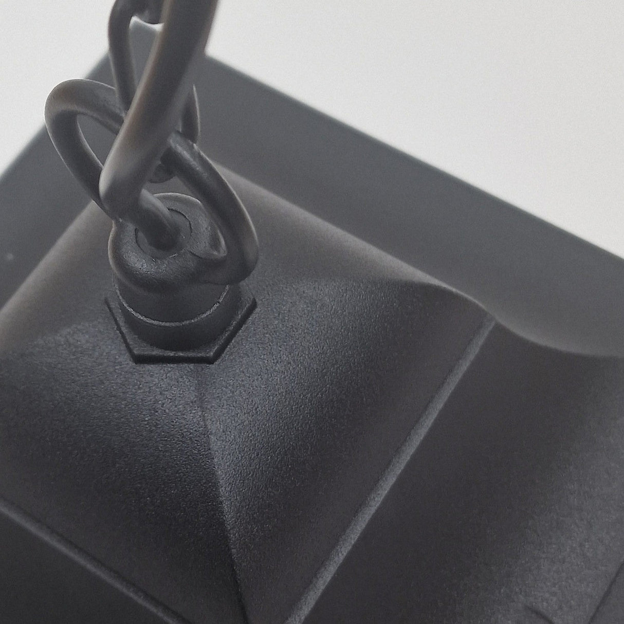 Close-up of a black metal chain connected to a textured, angular black mount, likely belonging to the Chapel 1 Light Outdoor Chain Pendant in die-cast aluminum. The background features a blurred, neutral white.
