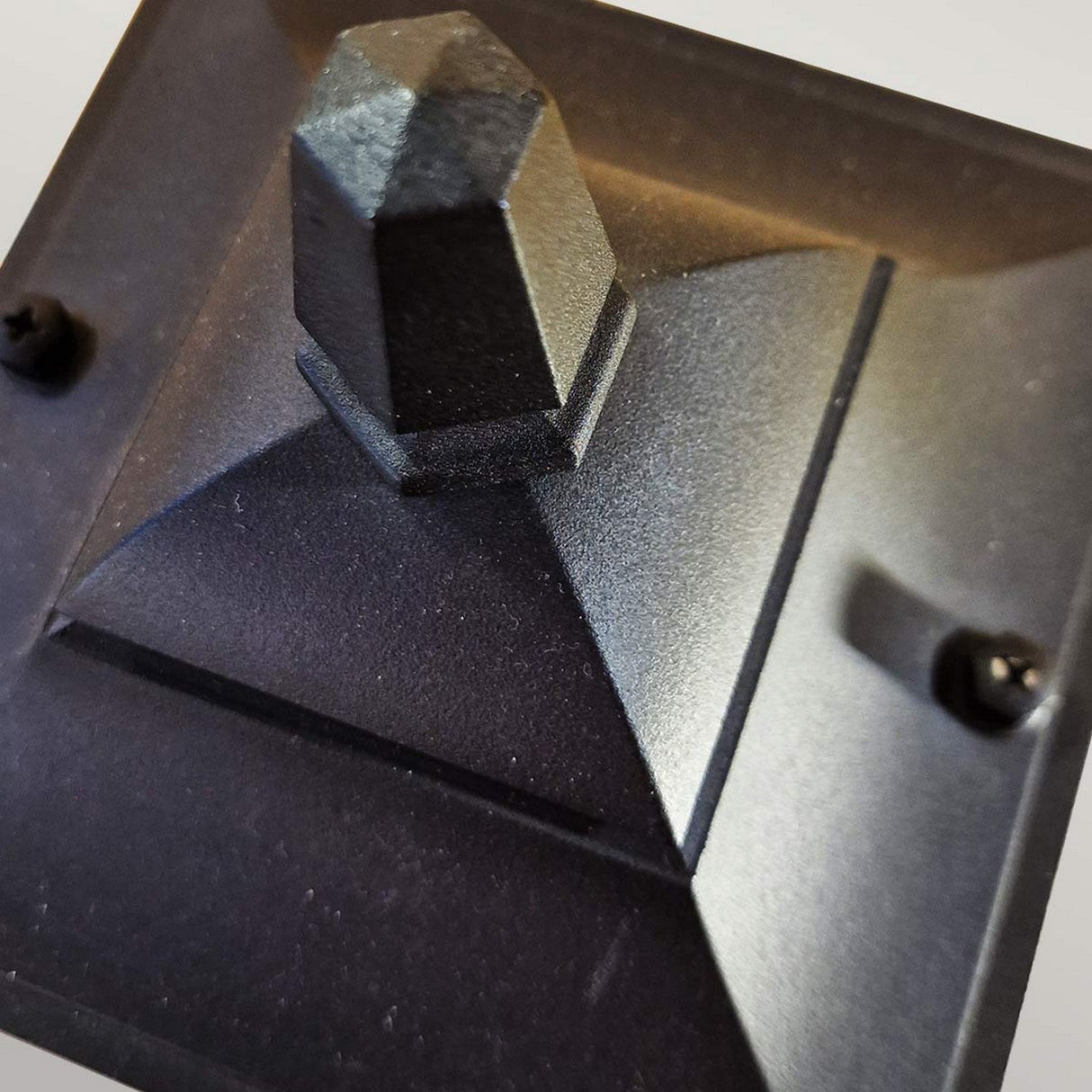 Close-up of the Chapel 1 Light Outdoor Mini Up Wall Lantern in black, showcasing a pyramid-shaped post cap crafted from die-cast aluminium with a hexagonal top. It has a textured surface with four visible screws at the corners, mounted on a square base.