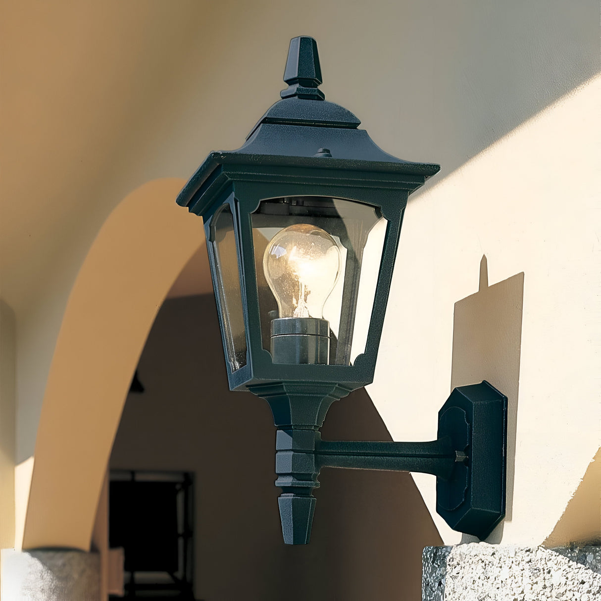 A black Chapel 1 Light Outdoor Mini Up Wall Lantern with a vintage design and visible bulb is mounted on a cream-colored wall. Its shadow plays against the surface, with an archway in the background. Crafted from die-cast aluminum, this elegant fixture adds timeless charm to any space.