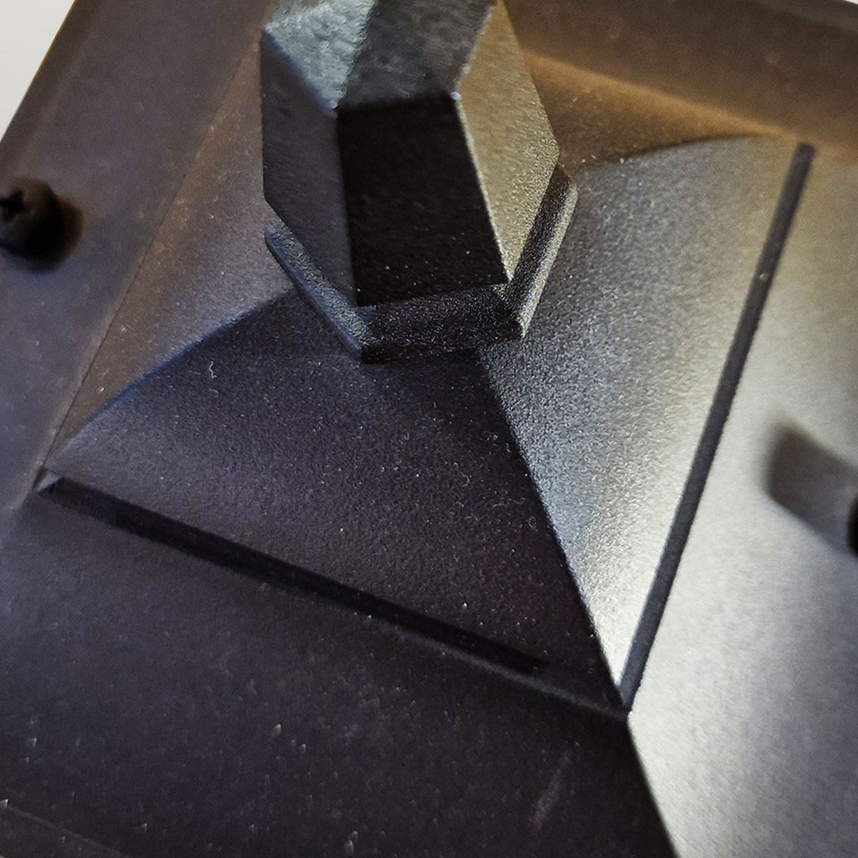 Close-up of the Chapel 1 Light Outdoor Mini Down Wall Lantern in black, showcasing its textured metallic pyramid shape crafted from die-cast aluminum. The perspective emphasizes its geometric design with sharp angles and a matte finish, reminiscent of modern outdoor lighting aesthetics.